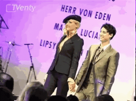 a man in a suit stands next to a woman in a beret holding hands in front of a sign that says herr von eden