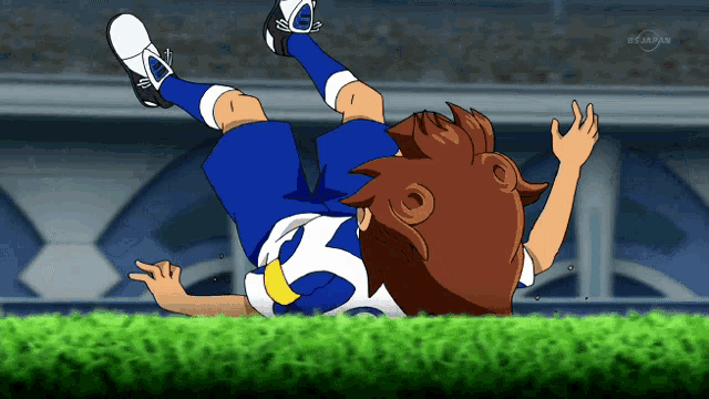a boy in a blue and white uniform is laying on his back on a soccer field with a tv behind him that says tokyo
