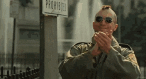 a man with a mohawk and sunglasses is clapping his hands in front of a prohibited sign .