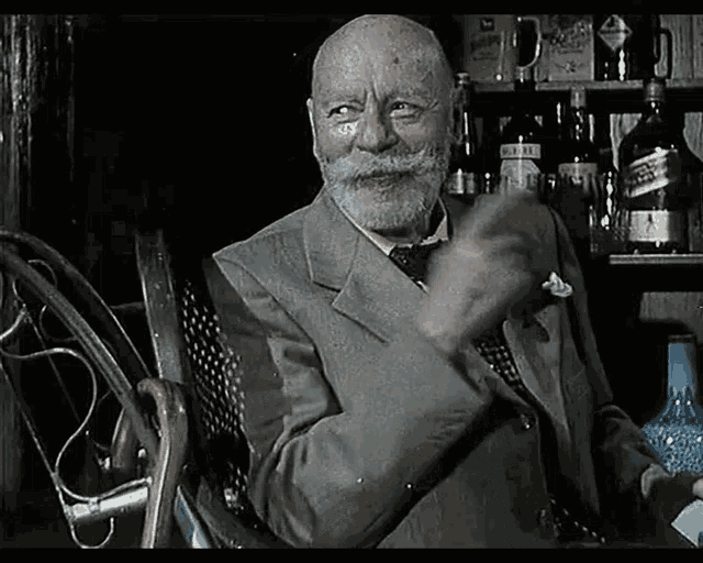 a man with a beard is sitting in a chair in front of a shelf with bottles of alcohol including johnnie walker