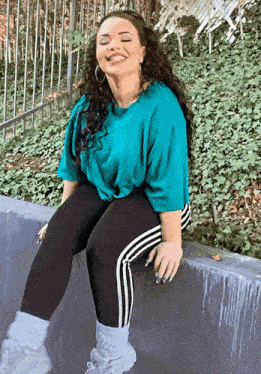 a woman wearing a blue shirt and black pants is sitting on a wall