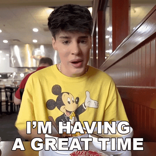 a young man wearing a yellow mickey mouse shirt says i 'm having a great time while sitting at a table