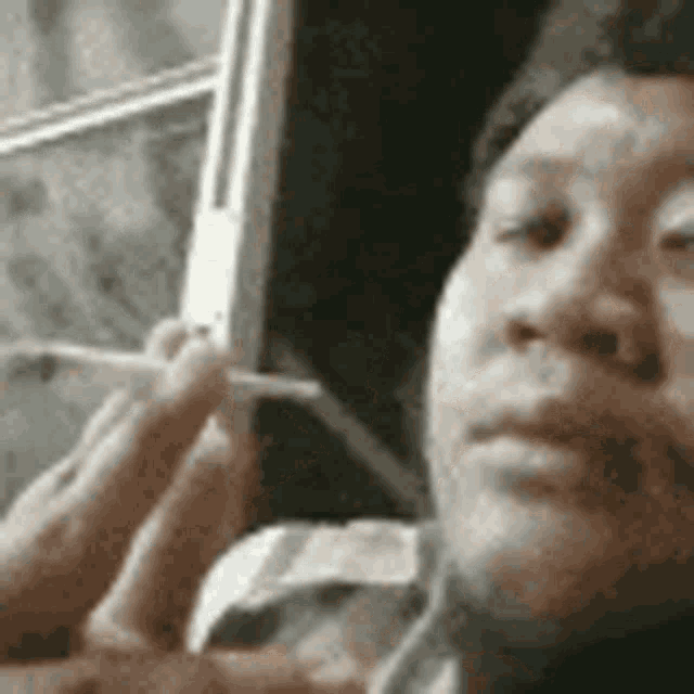 a man is smoking a cigarette in front of a window while looking at his phone .