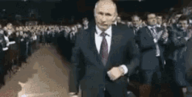 a man in a suit and tie is standing in front of a crowd of people applauding .
