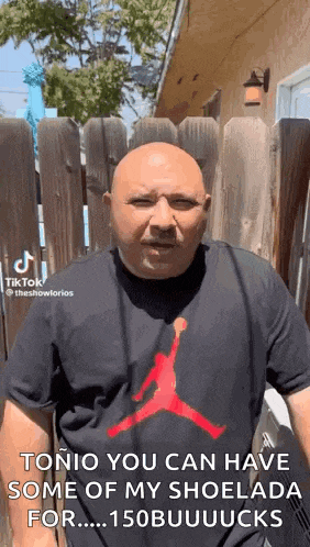 a bald man wearing a black shirt with a red air jordan logo is standing in front of a fence .