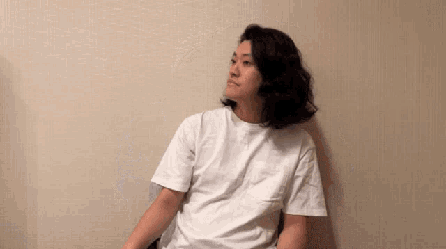 a man with long hair wearing a white shirt is sitting in front of a wall