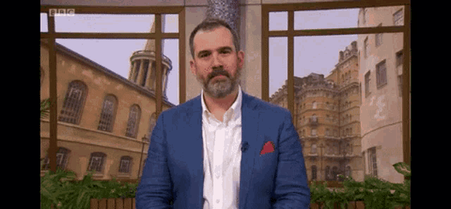 a man in a blue suit and white shirt is standing in front of a window with bbc written on it