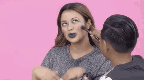 a man is applying makeup to a woman 's face with a brush on a pink background .