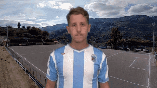 a man wearing a blue and white striped shirt stands in front of a soccer field with mountains in the background