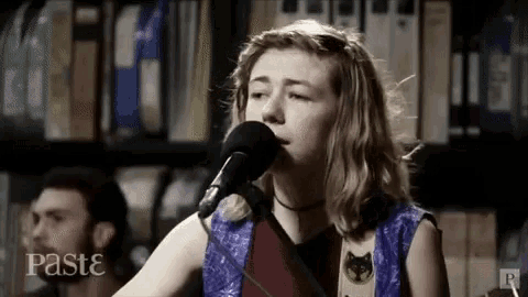 a young woman is singing into a microphone while playing a guitar in front of a bookshelf .