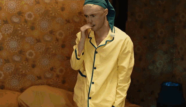 a man wearing a yellow pajama top and a green sleep cap stands in front of a bed
