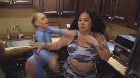 a group of women are fighting in a kitchen and one of them is wearing a blue shirt .