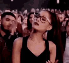 ariana grande is wearing glasses and a black dress while standing in front of a crowd .