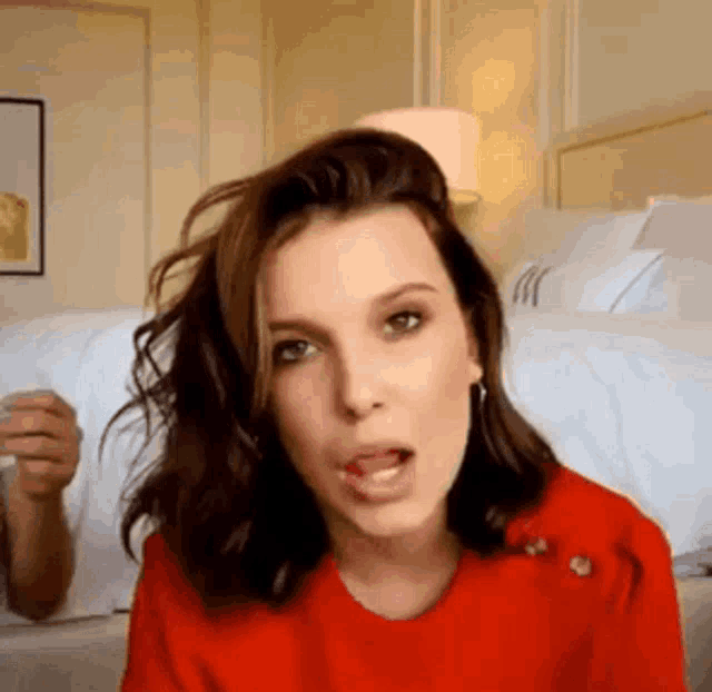 a woman in a red shirt is sticking her tongue out while sitting in front of a bed .