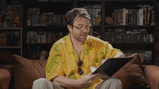 a man wearing glasses and a yellow shirt is sitting on a couch reading a book