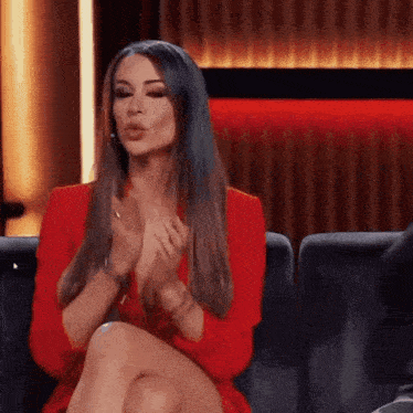 a woman in a red dress is sitting on a couch with her legs crossed and making a funny face .