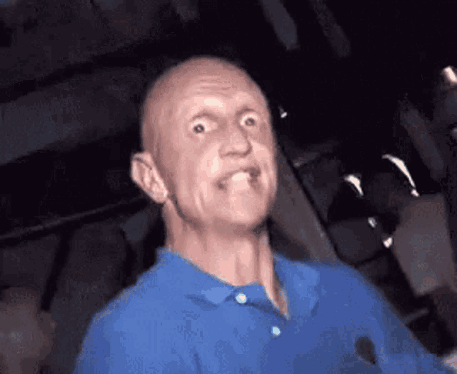 a bald man in a blue shirt is making a funny face in a dark room .