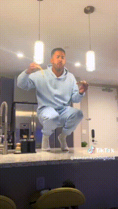 a man in a blue hoodie is squatting on a counter and eating a hamburger