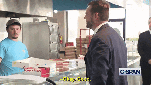 a man in a suit talks to a man in a blue shirt in a bakery