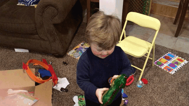 a little boy is playing with a green toy in front of a yellow folding chair