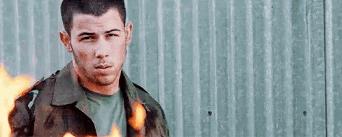 a man in a camouflage jacket is standing in front of a wall with fire coming out of his jacket .