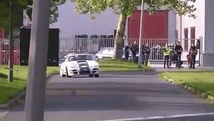 a white car is driving down a street in front of a group of people .