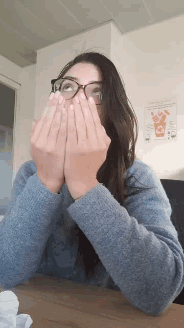 a woman wearing glasses and a blue sweater is covering her face with her hands