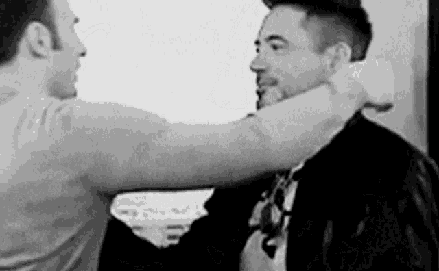 a black and white photo of two men hugging each other . one of the men is wearing a suit and tie .