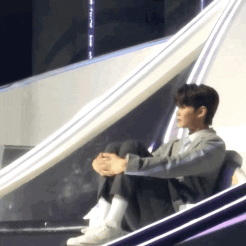 a man sits on a stage with his legs crossed and his hands folded