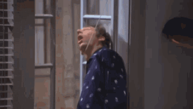a man in a blue pajama shirt is standing in front of a window with his mouth open .