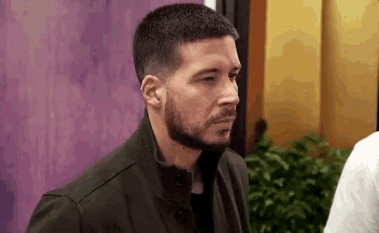 a man with a beard is standing in front of a purple wall and looking at something .