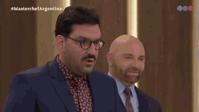 a man in a suit and tie stands next to another man in a suit and tie with the hashtag #masterchefargentina on the screen