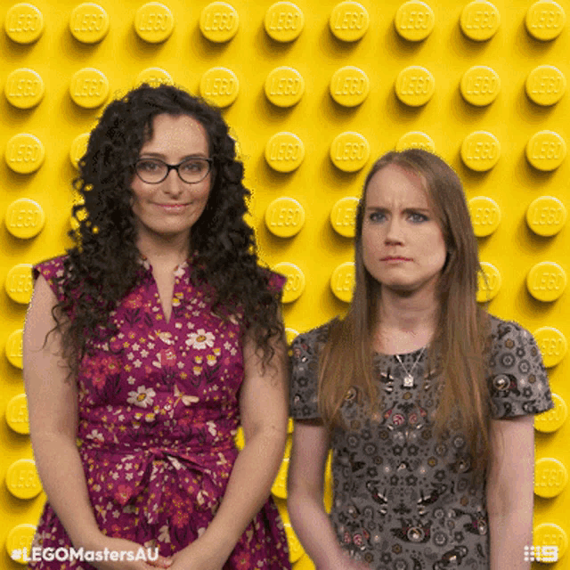 two women standing in front of a yellow wall with lego bricks on it