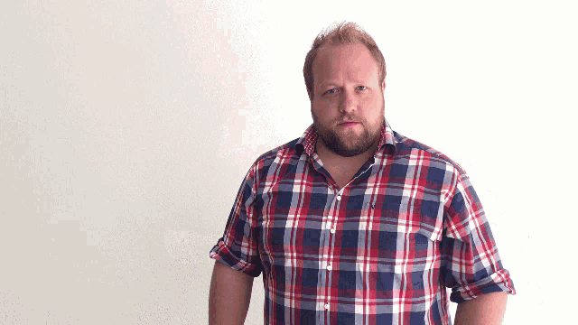 a man with a beard wearing a plaid shirt stands in front of a white wall