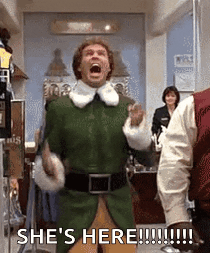 a man in a green elf costume is screaming and saying she 's here !!!