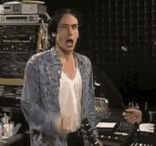 a man in a denim shirt and white tank top is holding a guitar and making a funny face .