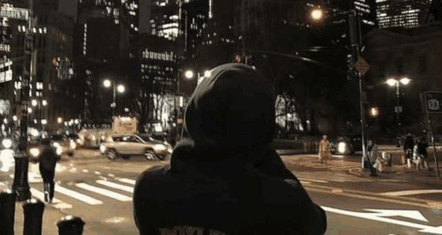 a person in a hoodie is standing on a city street at night looking at traffic .