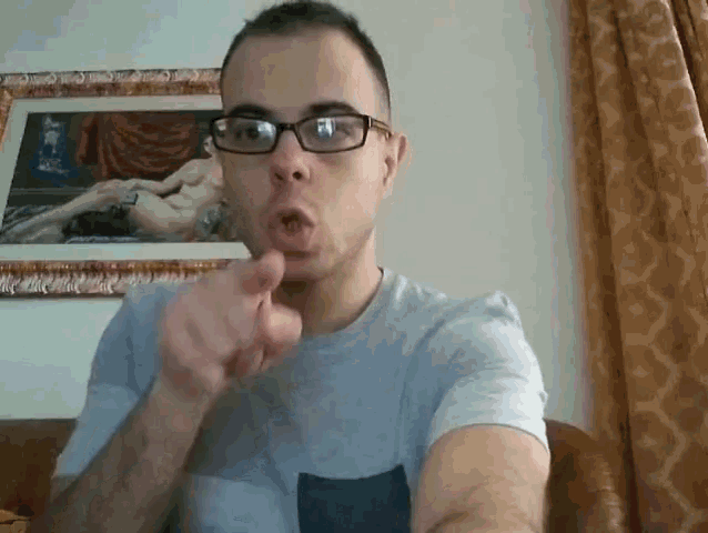 a man wearing glasses is pointing at the camera in front of a framed painting