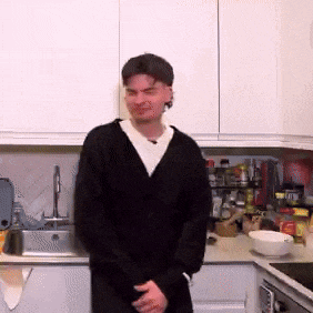 a man in a black cardigan stands in a kitchen
