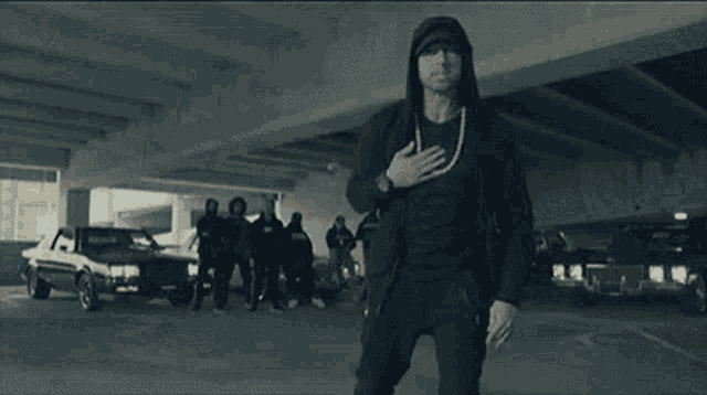 a man wearing a hooded jacket and a necklace is standing in a parking garage with cars .