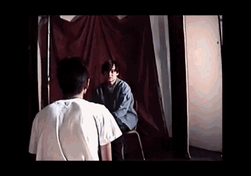 a man in a white shirt is standing next to a man in a chair in front of a red curtain .