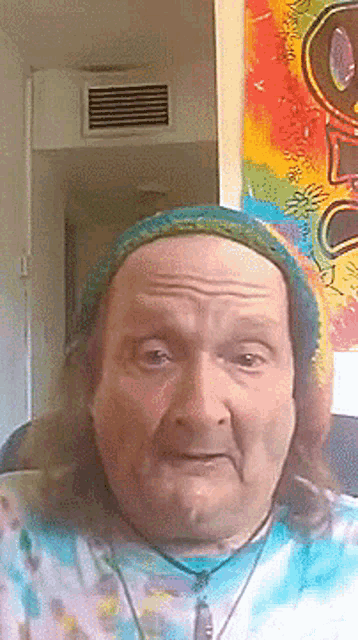 a man wearing a hat and a tie dye shirt is making a funny face .