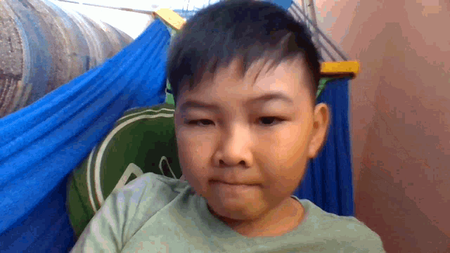 a young boy is sitting in a hammock with a green pillow and a blue curtain