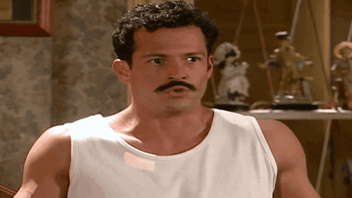 a man with a mustache wearing a white tank top looks at the camera