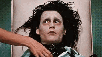 a man with long black hair is laying on a bed with a woman 's hand on his chest .
