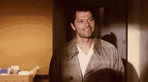 a man wearing a trench coat and a white shirt is smiling in a hallway .