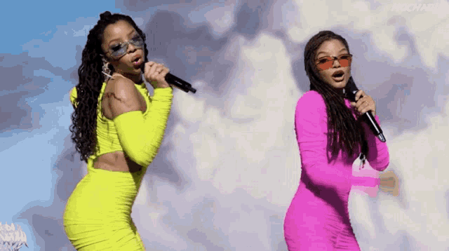 two women singing into microphones one in a neon yellow dress and the other in a bright pink dress