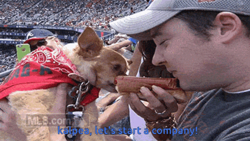 a man is eating a hot dog with a dog wearing a bandana next to him
