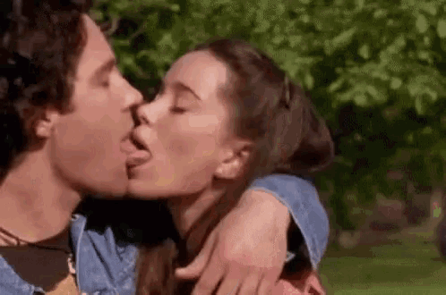 a man and a woman are kissing in a park . the woman is sticking her tongue out .