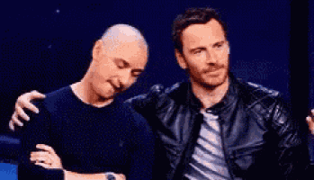 two men are sitting next to each other on a stage . one of the men has a bald head .